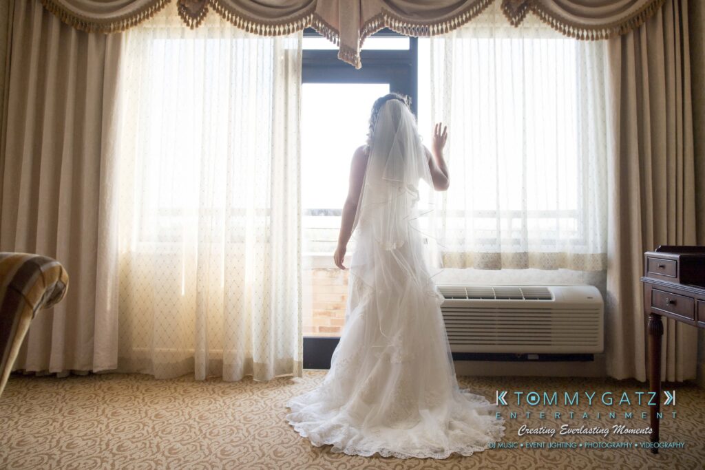 Bride in a white gown and veil stands by a window with sheer curtains, looking outside. Room has carpet flooring and an armchair.
