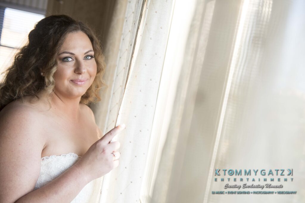 Bride in a strapless dress stands by a window, partially pulling back a sheer curtain. Logo for "Tommy Gatz Entertainment" is visible in the corner.