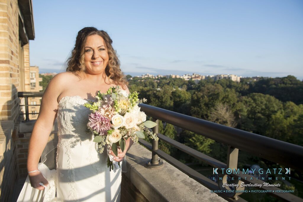 A bride in a wedding dress holds a bouquet while standing on a balcony with a scenic view of trees and a cityscape in the background.