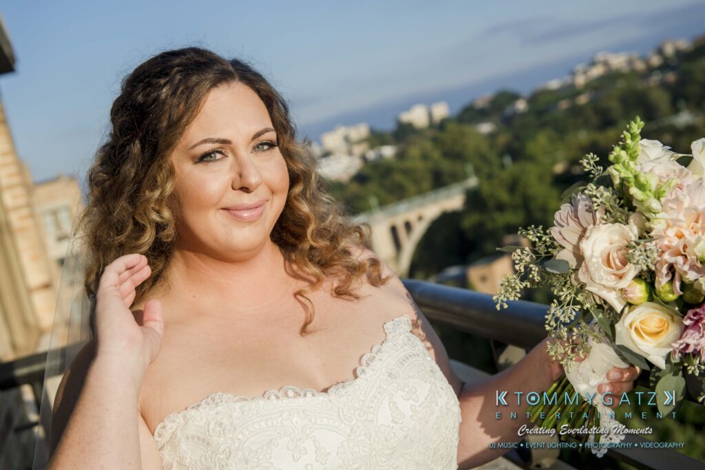 Bride in a white lace strapless dress holding a bouquet, standing on a balcony with a scenic view in the background.