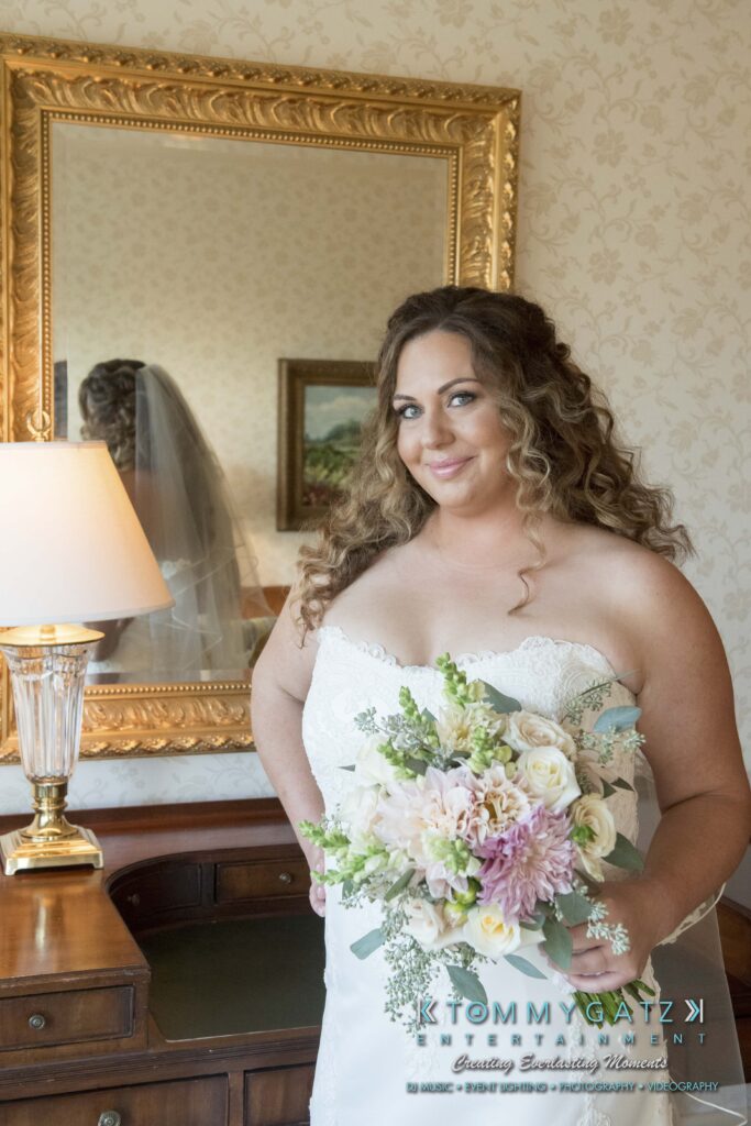 Bride smiling, holding a bouquet of flowers in a room with ornate wallpaper and a decorative mirror behind her. She wears a strapless dress and her hair is styled in loose curls.