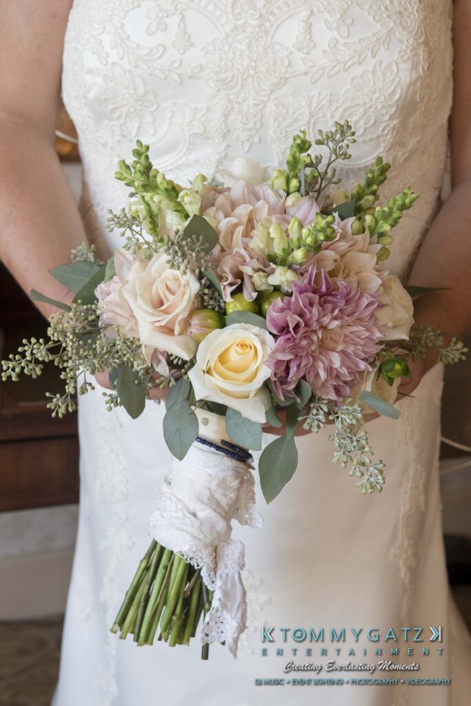 Bride holding a bouquet of roses, dahlias, and eucalyptus, with a lace-wrapped stem. She is wearing a white lace dress.