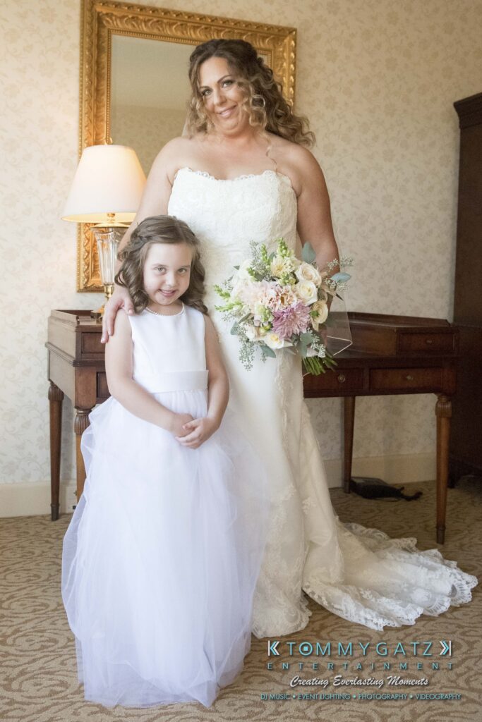 A bride in a white dress stands with a young girl in a white dress, holding a bouquet. They're indoors, in front of a mirror and a wooden dresser with a lamp.