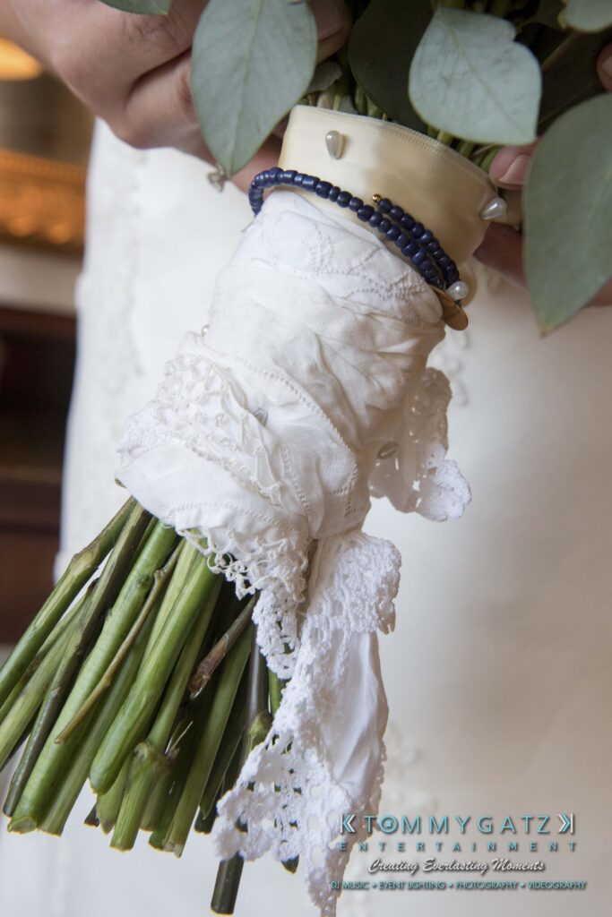 Close-up of a bride holding a bouquet wrapped in a lace cloth with a beaded bracelet and a small charm.