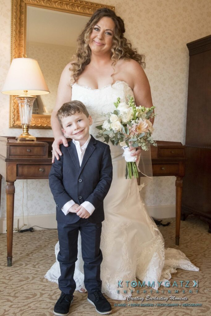 A woman in a white wedding dress holds a bouquet while standing beside a young boy in a suit. They are indoors, in front of a wooden dresser and mirror.