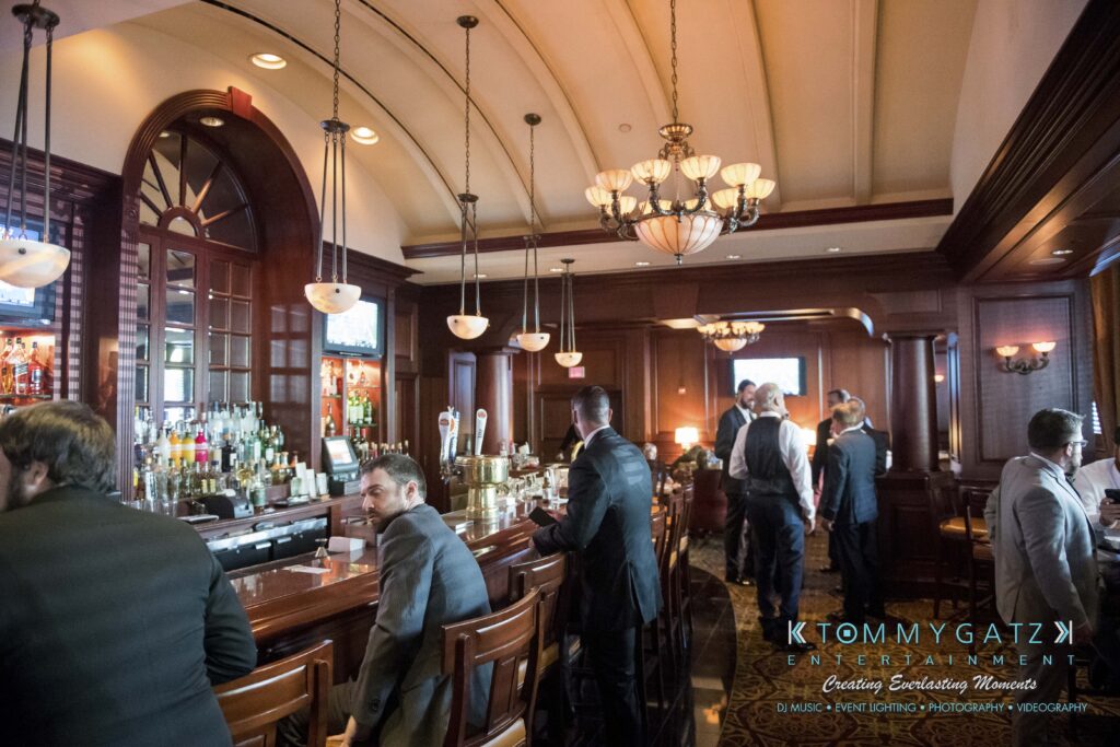A group of people in formal attire gather around a bar in a dimly lit room with wooden decor and chandeliers.