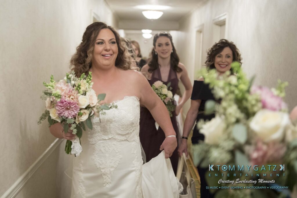A bride in a white dress walks with a bouquet, accompanied by smiling women in an indoor corridor.