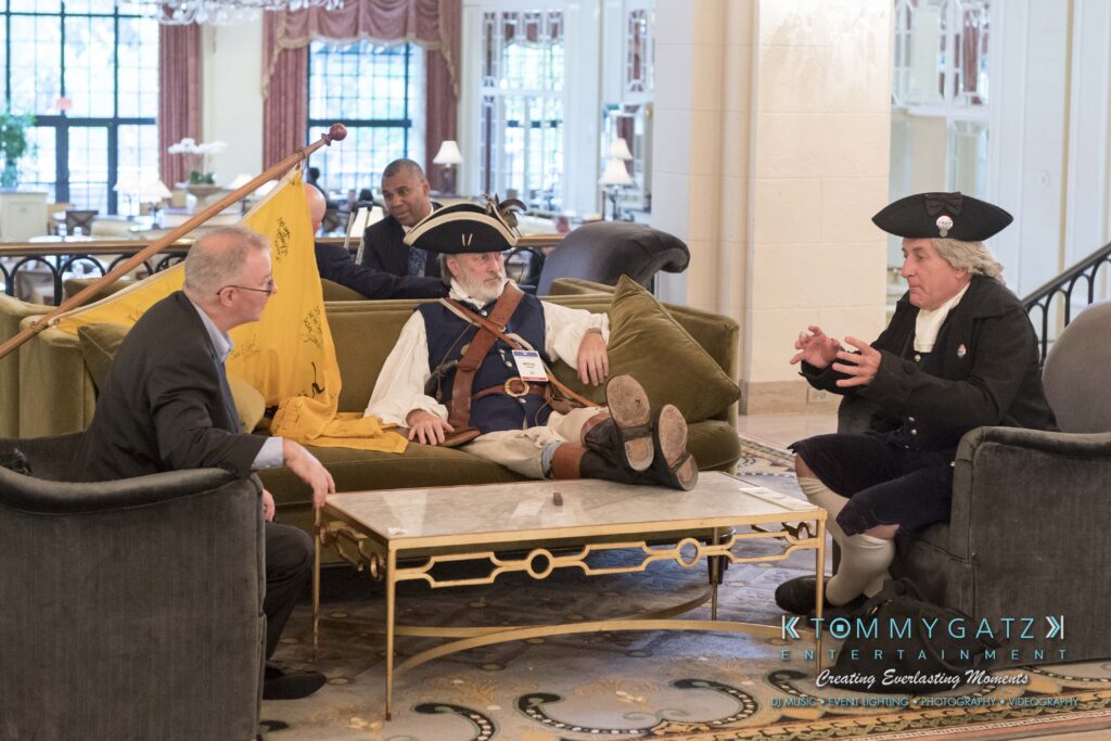 Three men in a hotel lobby, two in historical costumes with tri-corner hats, one in a suit. They're engaged in conversation, with one man reclining on a couch and a flag in the background.