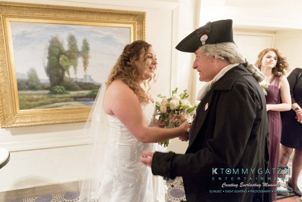A bride in a white dress smiles while shaking hands with a man dressed in colonial attire, next to a large framed painting.