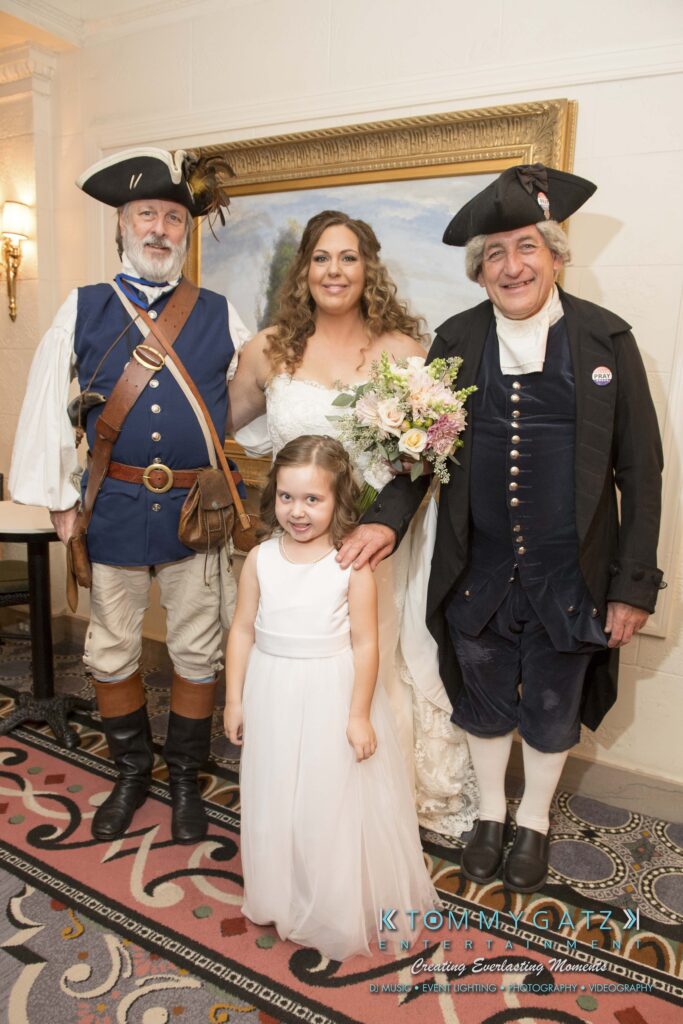 Bride holding a bouquet stands with a young girl and two men dressed in historical costumes.
