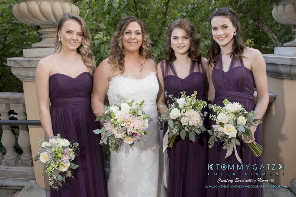 Four women in purple dresses stand next to a bride in white holding bouquets, posing outdoors.