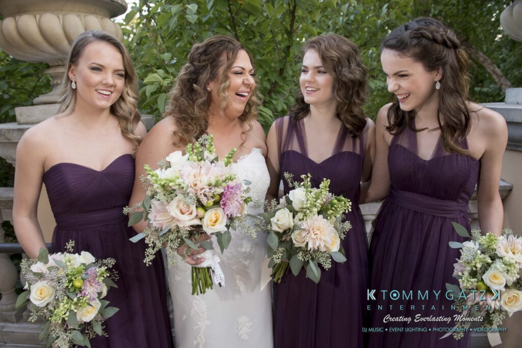 Four women in formal dresses, one in white holding a bouquet, stand together outdoors, smiling and conversing.