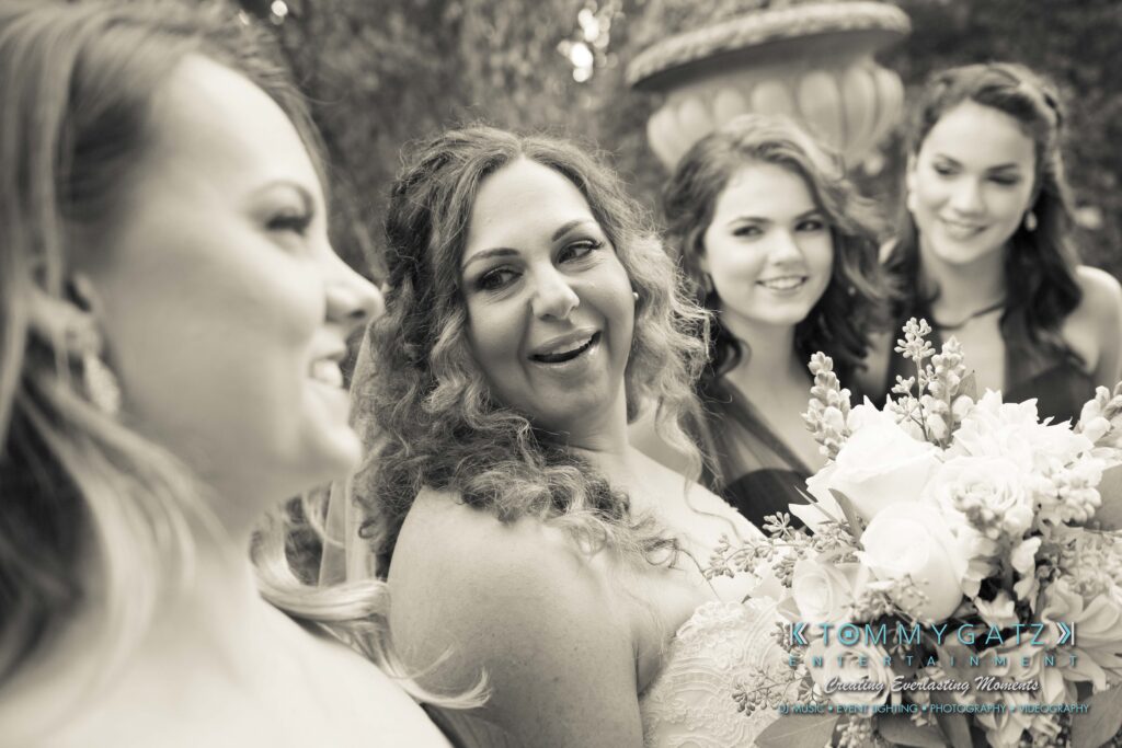 Four women in formal attire stand outdoors, three smiling and one holding a bouquet.