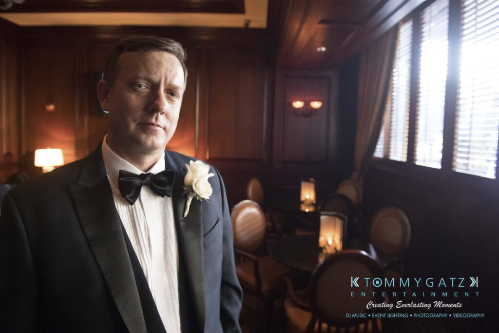 A man in a tuxedo with a white rose boutonniere stands in a dimly lit room with wooden walls and round tables. Text in the corner reads "Tommy Gatz Entertainment.