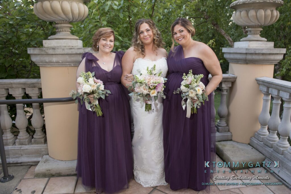A bride stands between two bridesmaids on a terrace. All are holding bouquets, and the bridesmaids wear matching purple dresses.
