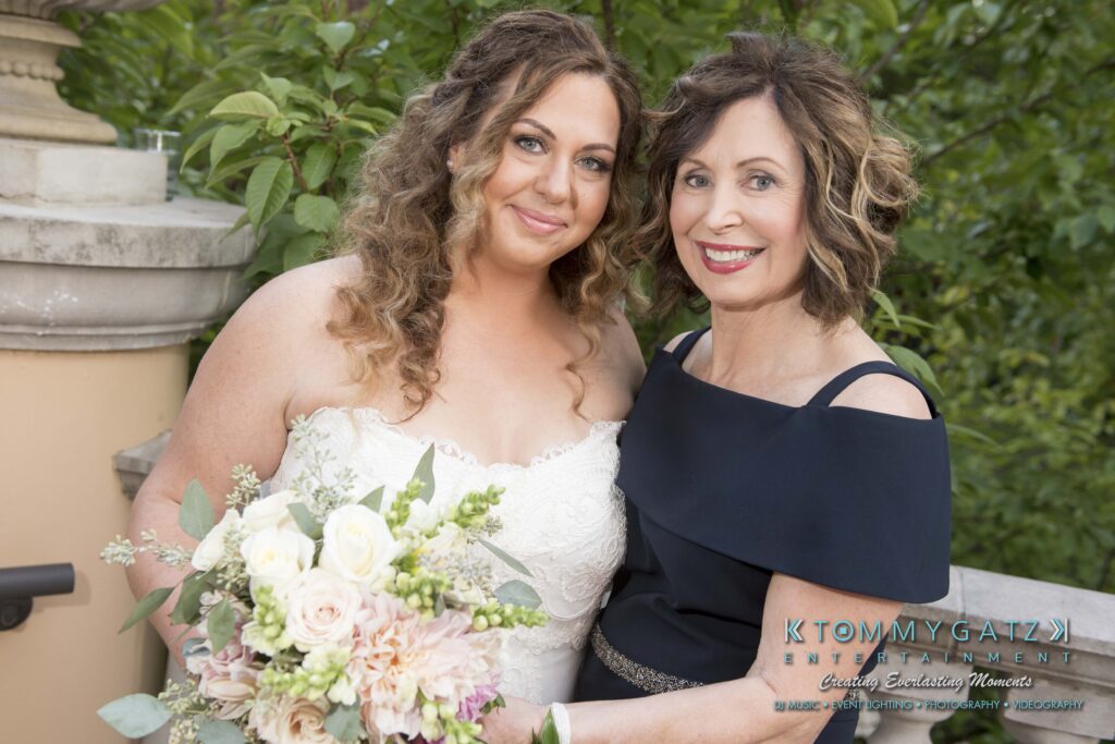 A bride in a white dress holds a bouquet while standing next to another woman in a dark dress. They are outdoors with greenery in the background.