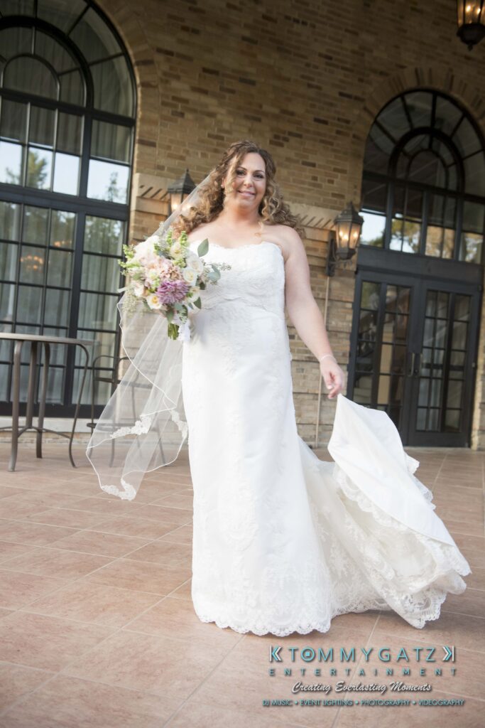 A bride in a white dress and veil holds a bouquet while standing on a patio. She is smiling and holding her dress with one hand. The background features arched windows and brickwork.