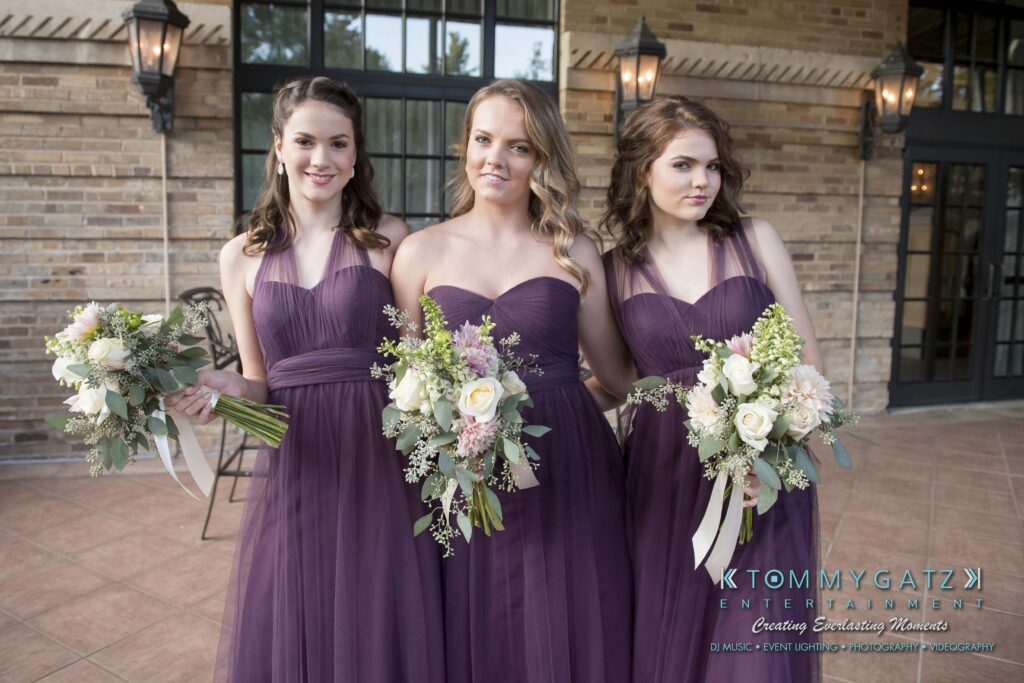 Three women in matching purple dresses hold bouquets while standing in front of a brick wall.