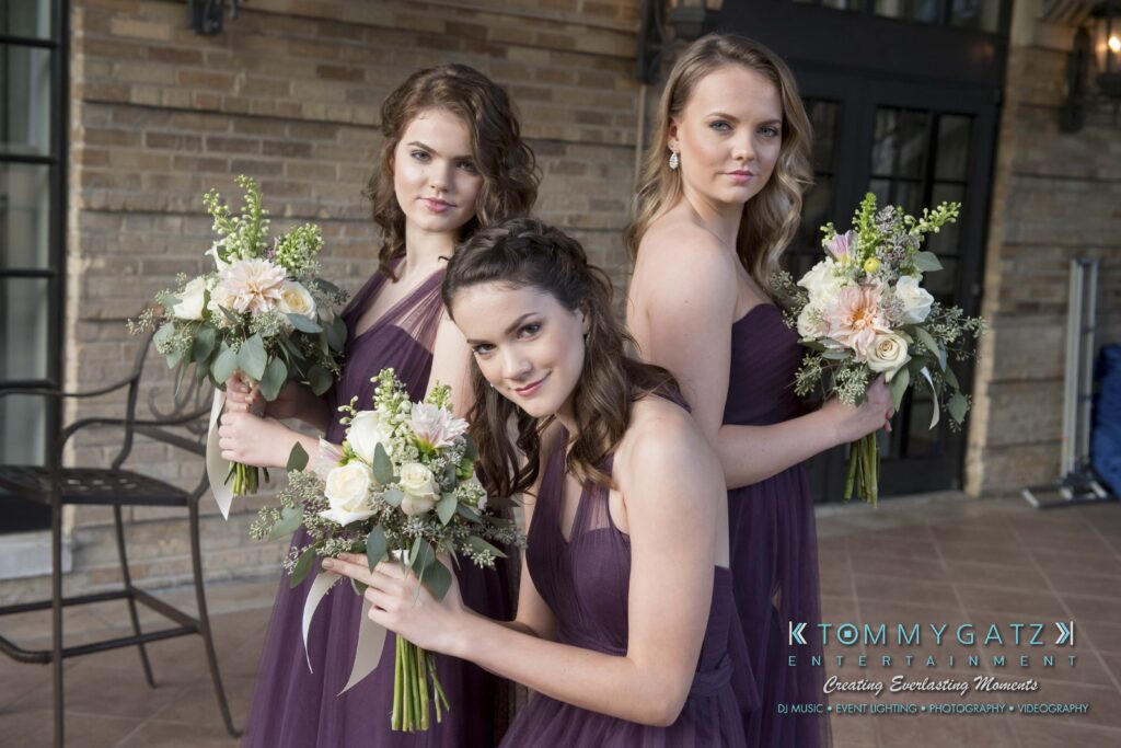 Three women in purple dresses holding bouquets pose together indoors.