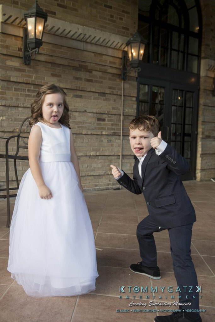 Two children outdoors; a girl in a white dress stands with a calm expression, while a boy in a suit strikes a playful, energetic pose. A brick building wall and lanterns are in the background.