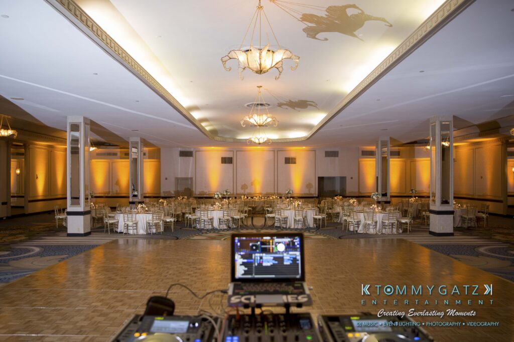 Empty ballroom set up for an event with round tables and chairs, a DJ booth in the foreground, and chandeliers hanging from the ceiling.