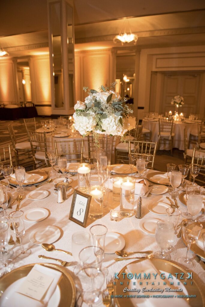 Elegant table setting with white floral centerpiece, candles, glasses, and cutlery on a round table. A table number card is displayed. Warm lighting in a banquet hall.