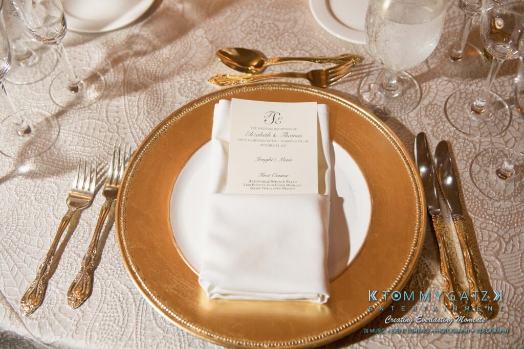 Elegant table setting with a gold-rimmed plate, gold cutlery, and a folded napkin holding a printed menu card. Crystal glasses and a textured tablecloth complete the arrangement.