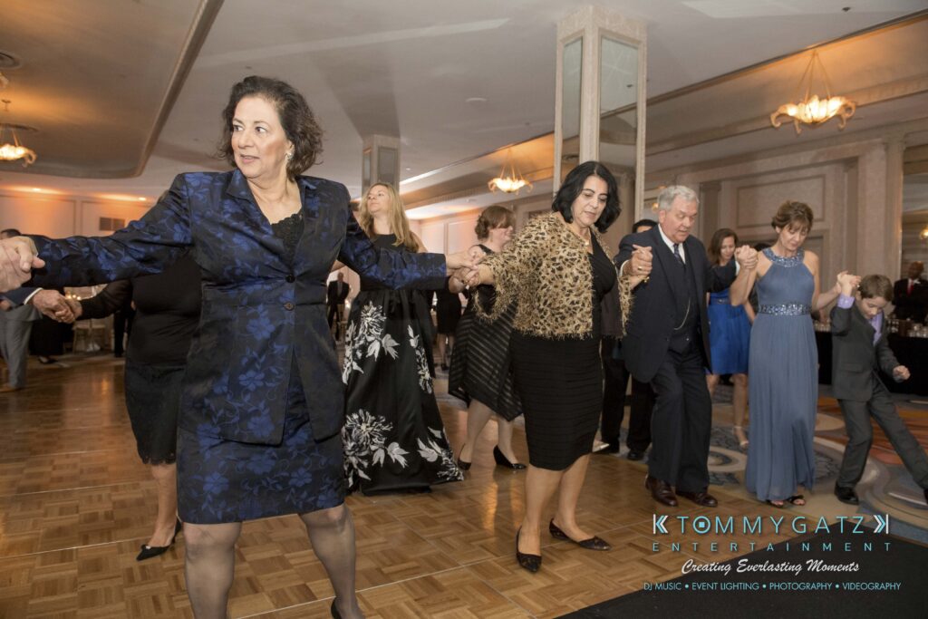 A group of people dancing in formal attire at an indoor event, holding hands in a line.