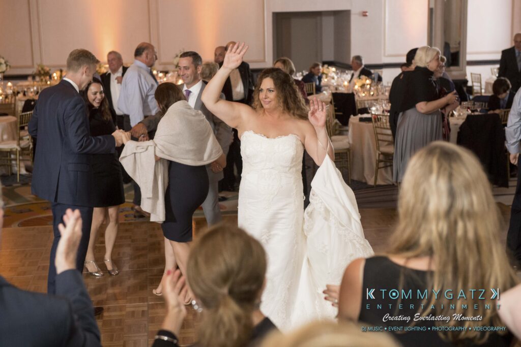 Bride in a white gown dancing with guests on a ballroom dance floor, surrounded by people in formal attire.