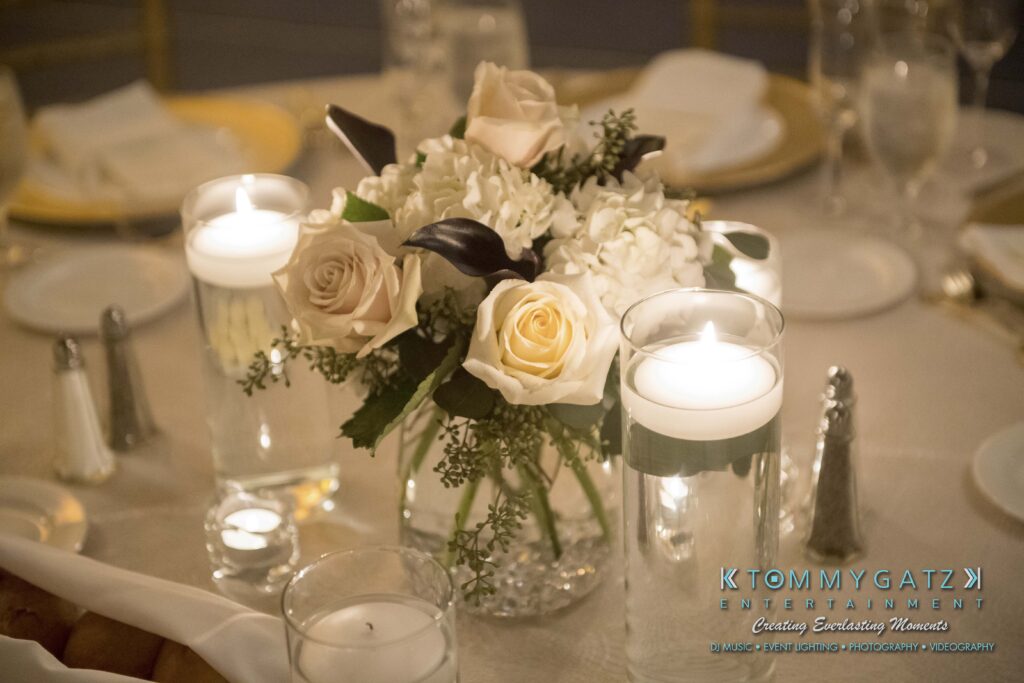 A table centerpiece with white and pale pink roses, hydrangeas, and candles in glass holders, surrounded by dinnerware.
