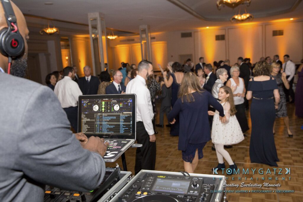 DJ playing music at a lively event with a large group of people dancing on a wooden floor in a warmly lit room.