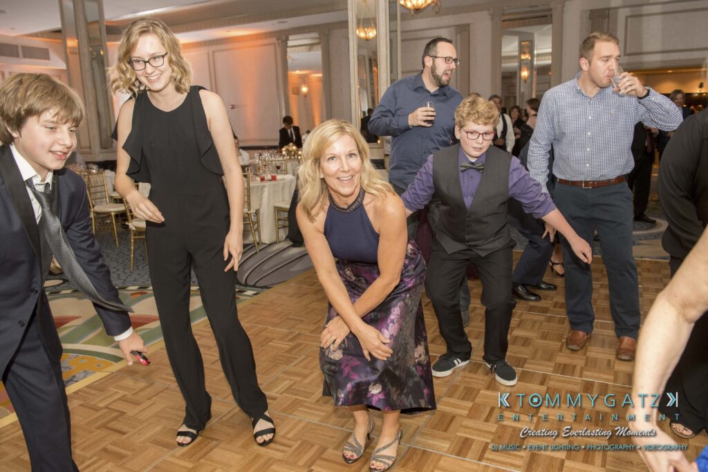 People in formal attire dancing in a brightly lit banquet hall with a wooden floor.