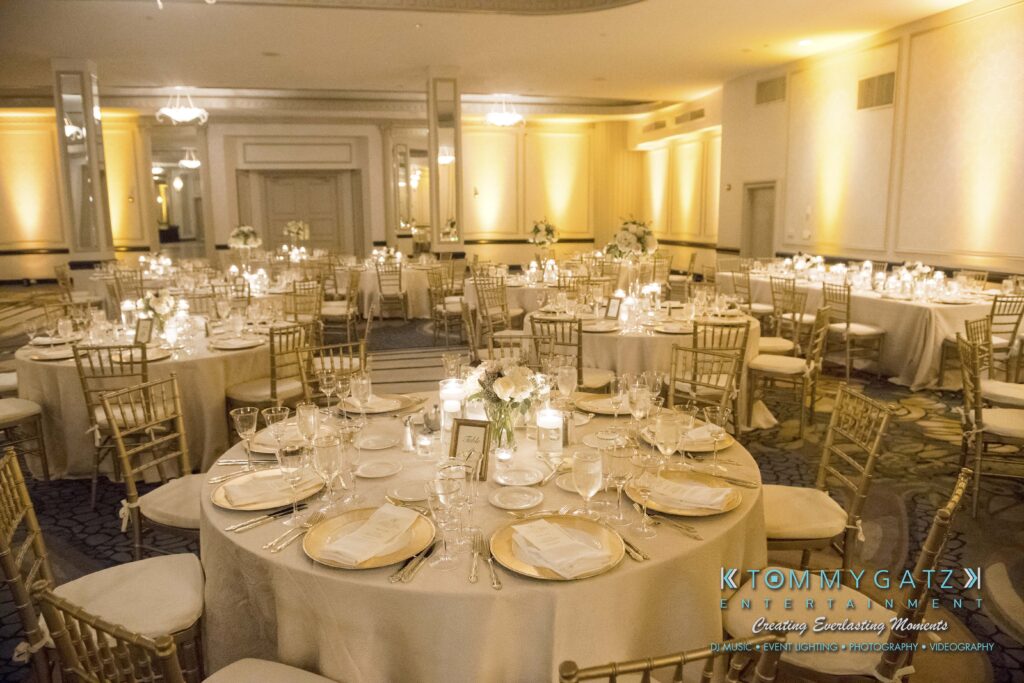 Elegant banquet hall with round tables set for a formal event. Each table is adorned with candles, white flowers, and neatly arranged dinnerware. Gold chairs surround the tables, and the room is softly lit.