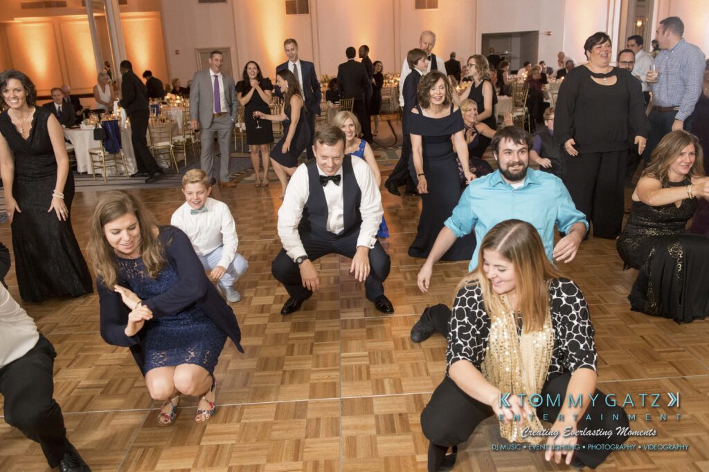 People on a dance floor participating in a group dance. Some are kneeling with hands out, while others are standing. Tables and other guests are visible in the background.