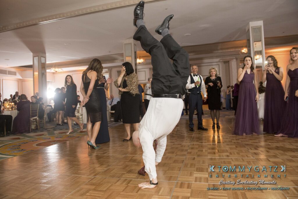 Man doing a handstand on a dance floor at an event, surrounded by people holding drinks and wearing formal attire.