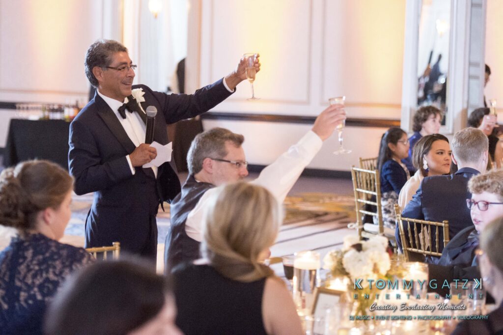 A man in a tuxedo gives a toast with a raised glass at a formal event. Guests seated at tables listen and raise their glasses in response.