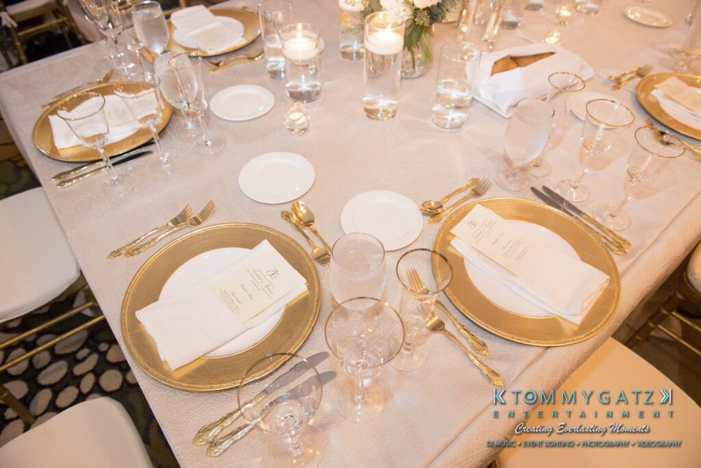 Elegant table setting with gold-rimmed plates, white napkins, and gold utensils on a white tablecloth. Glassware and small floral centerpieces are arranged neatly.