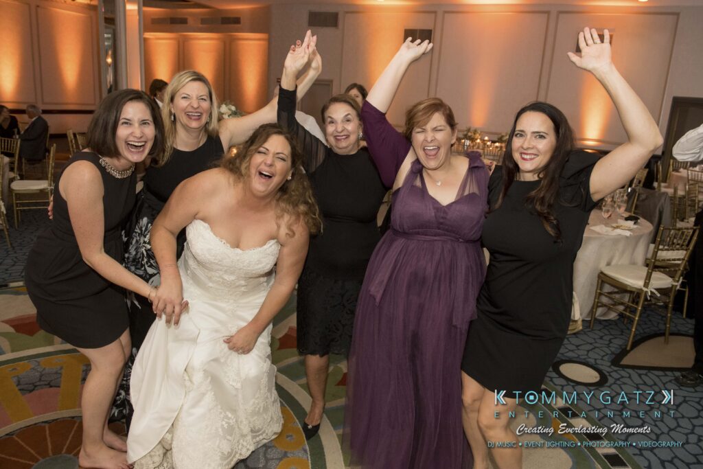 Bride and five women in a group pose, smiling and raising their arms, at an indoor wedding reception.
