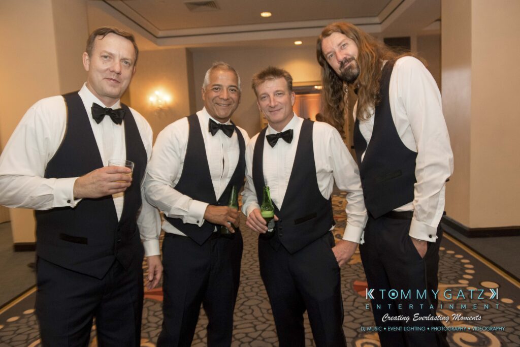 Four men in formal attire wearing vests and bow ties stand together, two holding drinks, in a carpeted indoor setting.