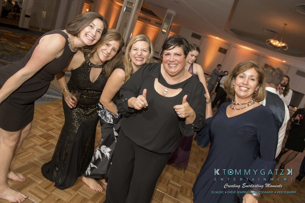 Five women in formal attire smile and pose on a wooden dance floor at an indoor event.