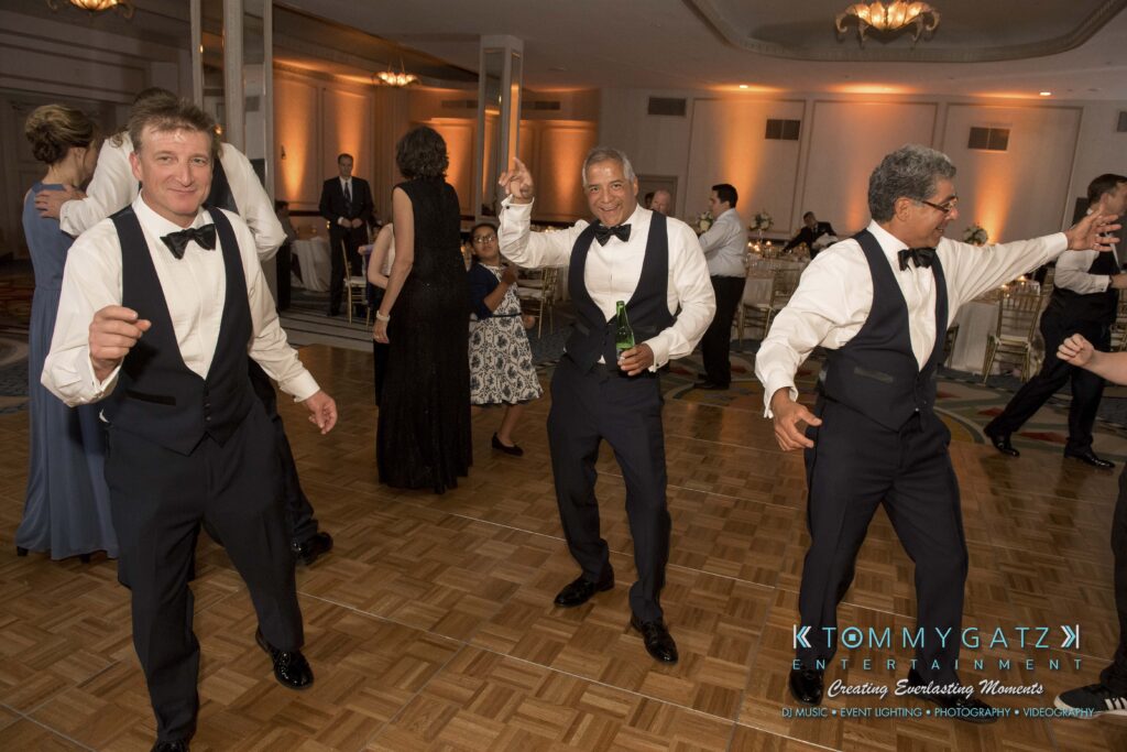 Three men in formal wear dance joyfully on a wooden dance floor at an indoor event. Other attendees and tables are visible in the background.