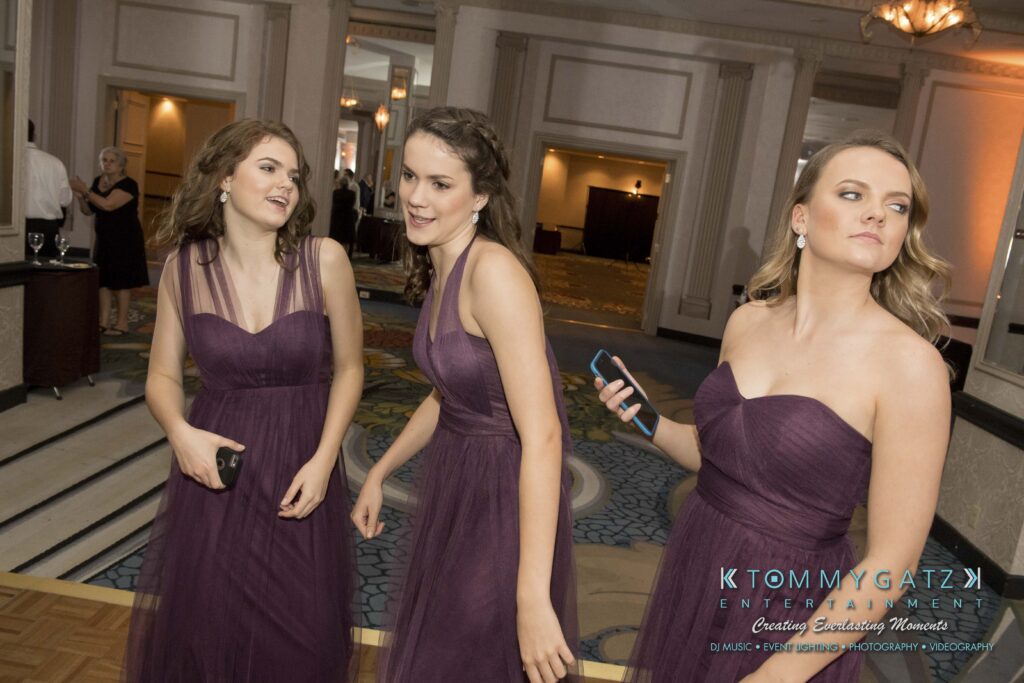 Three women in purple dresses stand in a formal venue, each holding a phone. They appear to be engaged in conversation, surrounded by decorative elements.