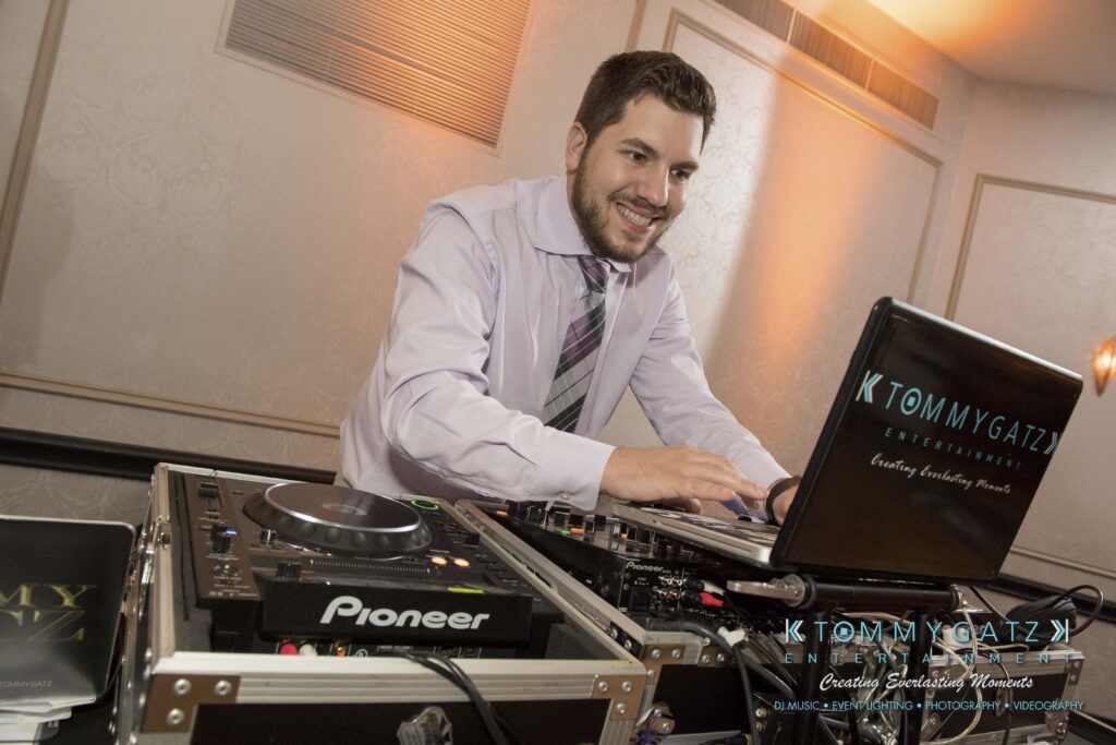 A DJ wearing a light purple shirt and patterned tie operates music equipment in an indoor setting.