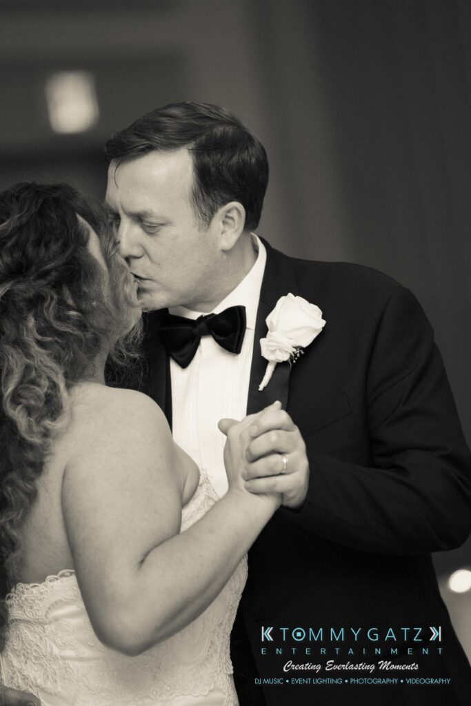 A couple dressed in wedding attire share a kiss on the dance floor. The groom wears a tuxedo with a bow tie and boutonnière, while the bride has curly hair and a strapless gown.
