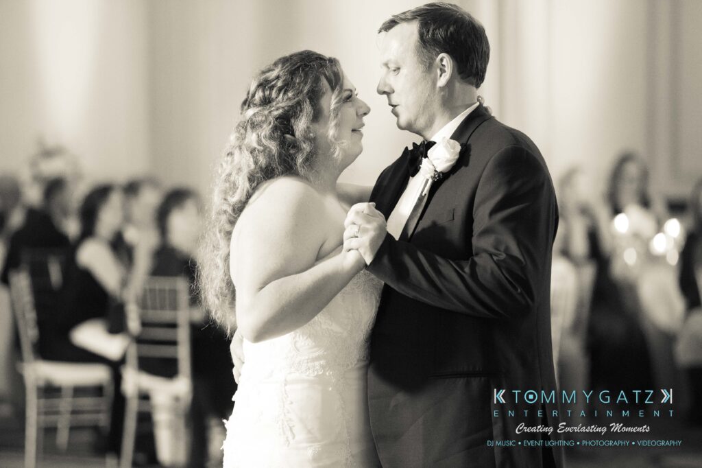 A couple dances closely at an event, surrounded by guests seated at tables.