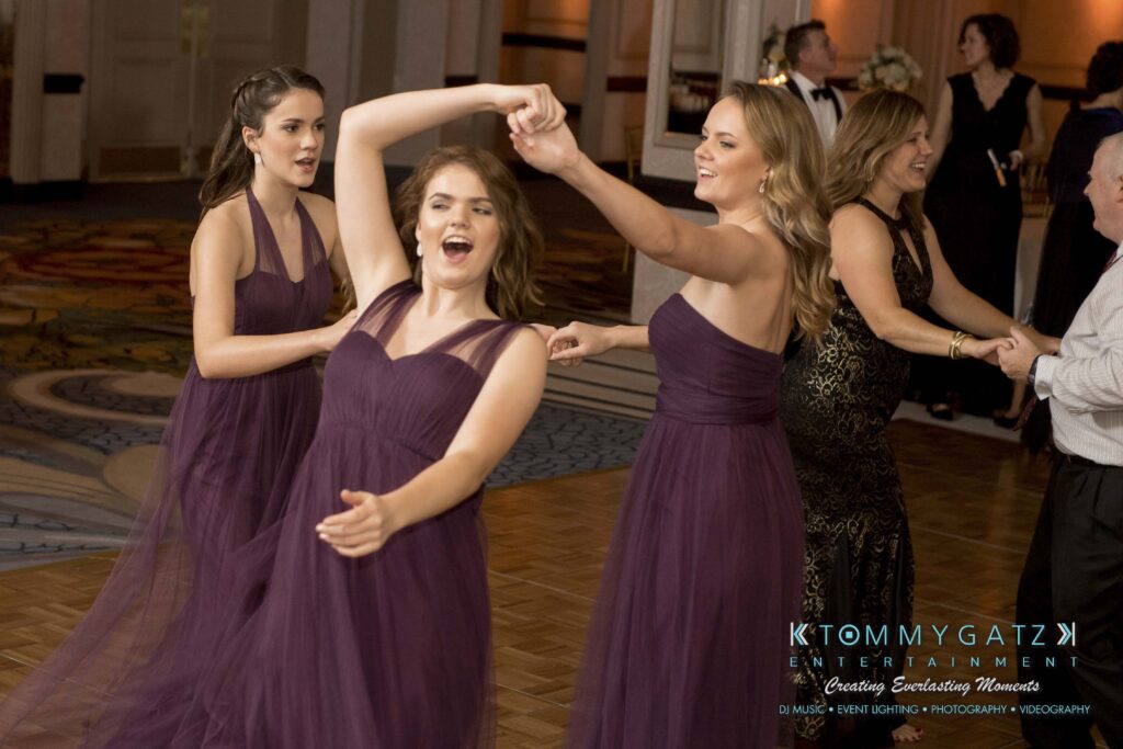Three women in purple dresses dance joyfully on a ballroom floor with other people in formal attire visible in the background.
