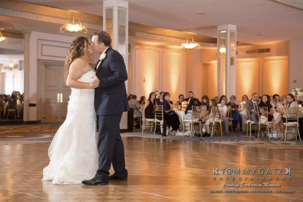 A bride and groom share their first dance in a warmly lit banquet hall, surrounded by seated guests.