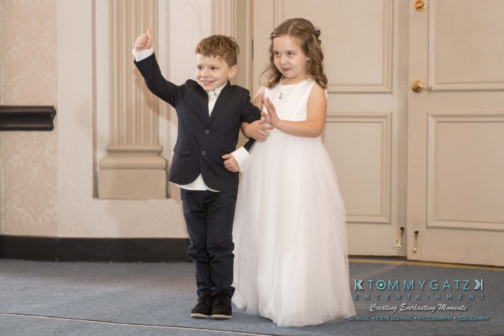 A young boy in a suit waves, standing alongside a girl in a white dress. They are indoors in front of closed double doors.