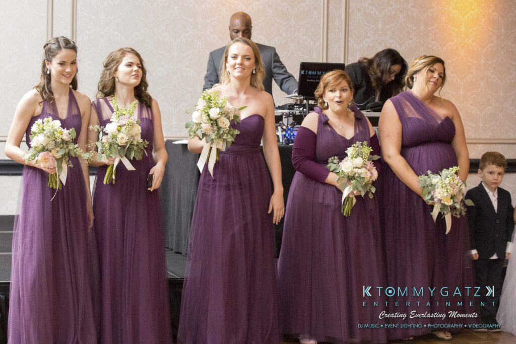 Five women in matching purple dresses and holding bouquets stand in a line at an event. A DJ and young boy are in the background.