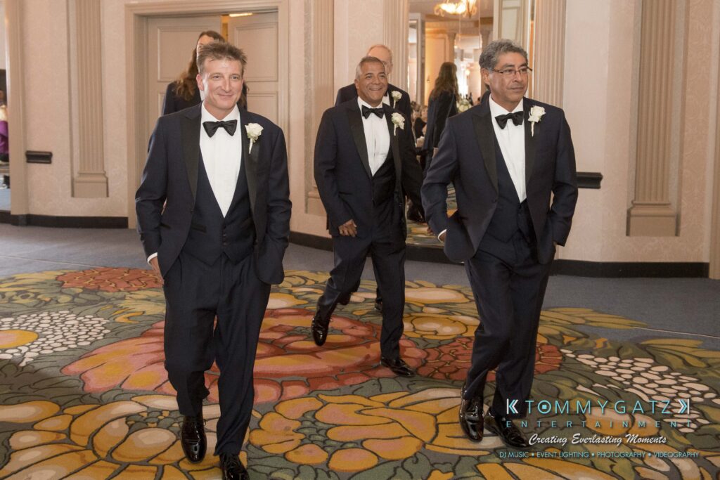 Three men in tuxedos walk through a carpeted ballroom, smiling. Large mirrors and beige walls are in the background.